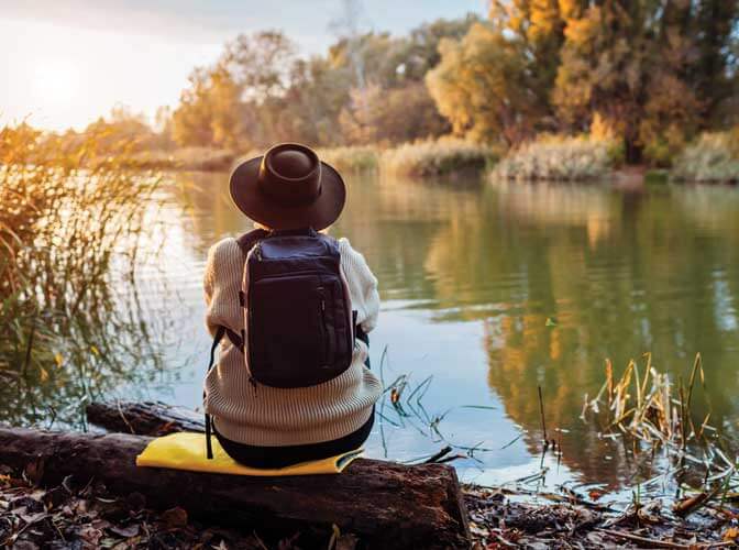 person with a backpack wearing a hat sitting on a log looking out over a lake