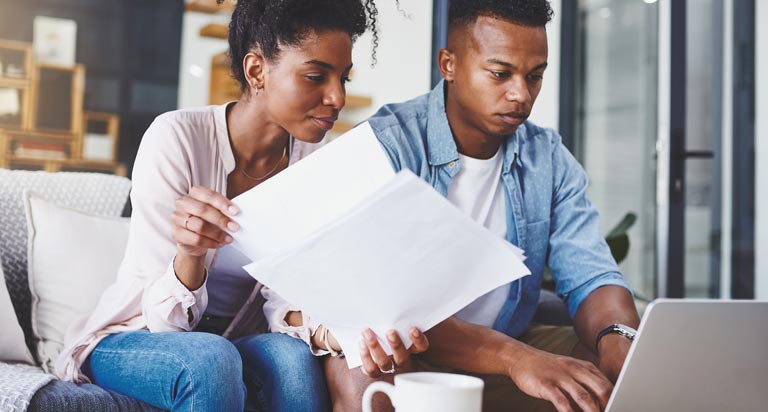 A young couple sits on a couch with documents in hand, looking down at a laptop in front of them with intensity.