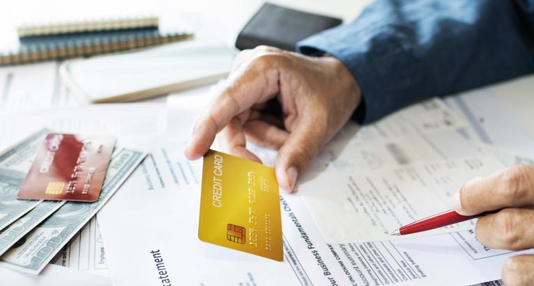 A surface covered in financial statements, a credit card and cash. A person’s arms reach over with a pen and credit card.