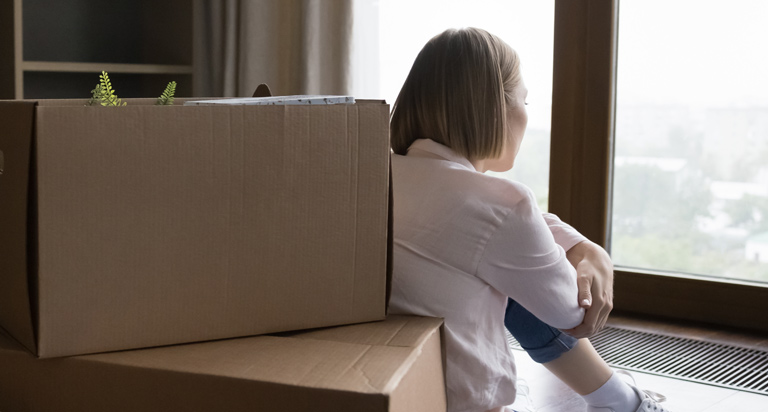 Stacks of cardboard moving boxes sit on the floor as a woman sits next to them staring pensively at a set of windows.