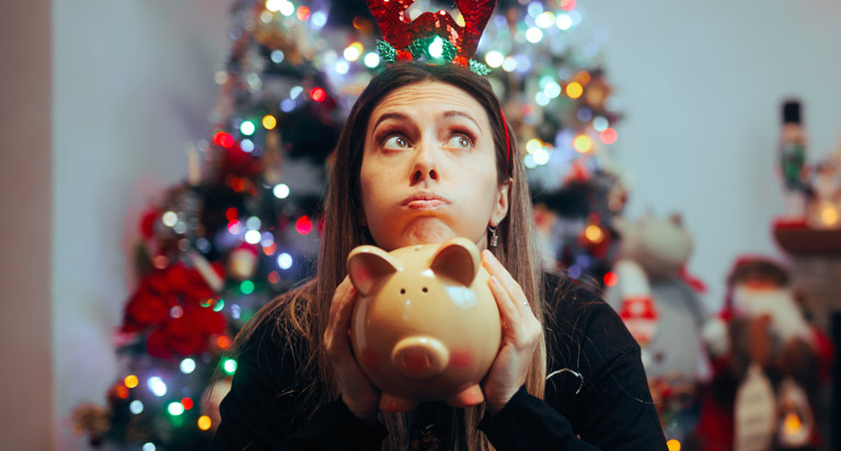 A quizzical looking woman sits in front of a Christmas tree and other holiday decorations with a piggy bank in her hands.
