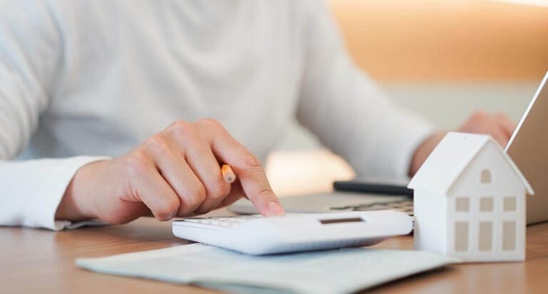 A woman taps on a calculator with her right hand and a computer with her left. A small plastic house in the foreground.