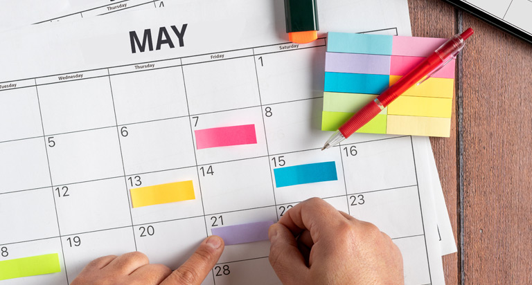 Three months of a calendar are spread across a table. A pair of hands use sticky notes to color-code certain days of the week.