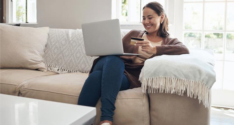 A smiling woman sits on a couch with her laptop on her knee and a credit card in her left hand.