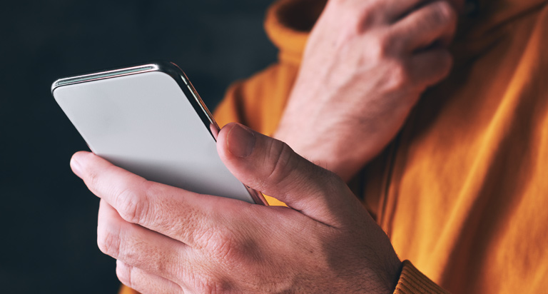 A man looks down at a phone he’s holding in one hand. He’s rubbing his bearded chin with the other hand.
