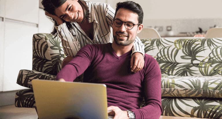 A smiling woman leaning on a man wearing glasses who is smiling while looking and pointing at his laptop