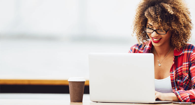 A woman wearing glasses that is smiling at her laptop that is sitting next to her coffee cup
