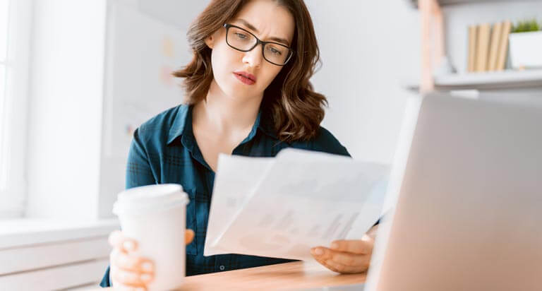 A woman in glasses sits in front of a computer holding a coffee cup in her right hand as she looks down at papers in her left hand.