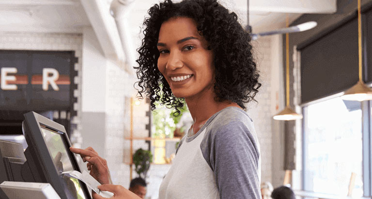 A woman stands in front of a point-of-sale computer and types on it with her right hand while smiling and holding papers in her left hand