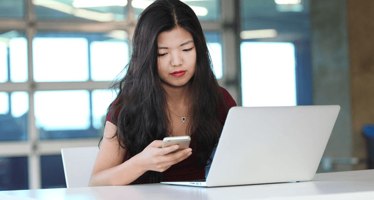 A woman looking down at her phone in her right hand while her laptop sits open in front of her