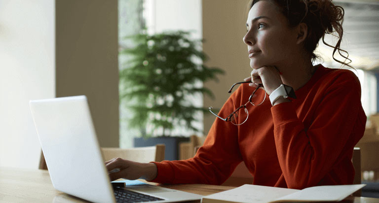 A woman rests her head on her left hand that is holding her glasses while she looks out a window and her right hand rests beside her opened laptop