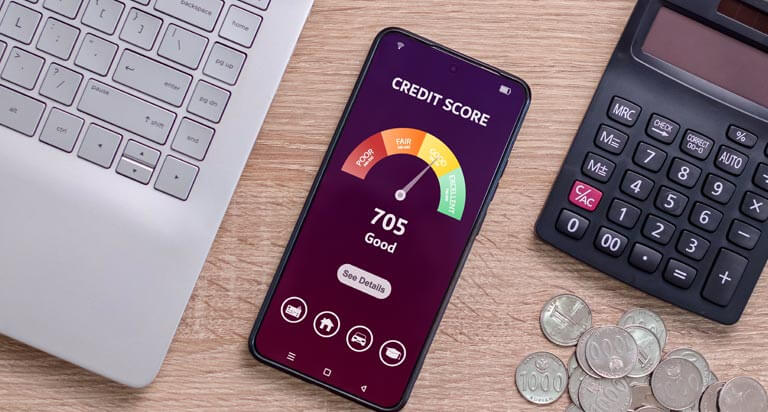 A mobile phone displaying a credit score gauge sits in the middle of a wooden desk surrounded by a computer, a calculator, and coins.