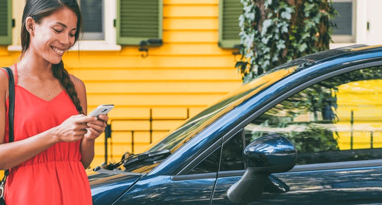 A woman who is standing next to her car and is looking happily at her phone in her hand