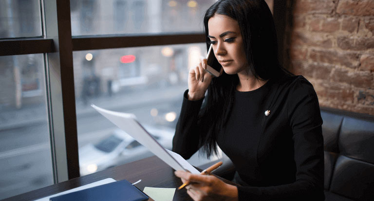 A woman holding a cellphone up to her ear as she looks at the papers in her left hand