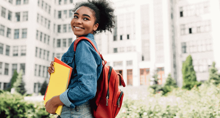 A woman wearing a read backpack and holding a red and yellow notebook looking over her shoulder and smiling