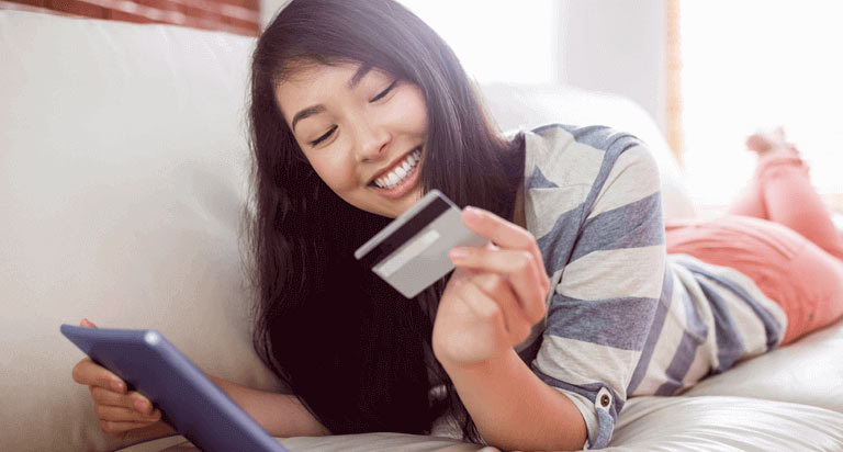 A woman looking down at a credit card in her left hand and smiling while holding a blue tablet in her right hand