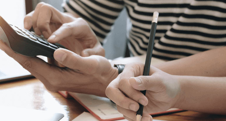 A person types on a calculator while someone else writes something with a pencil