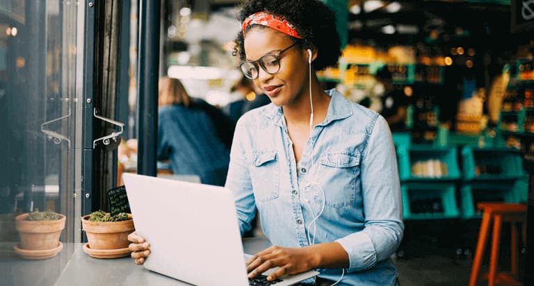 A woman wearing glasses and earbuds holding and looking at her laptop
