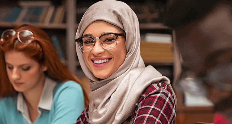 A woman wearing glasses and a hijab smiling and a woman in the background focusing