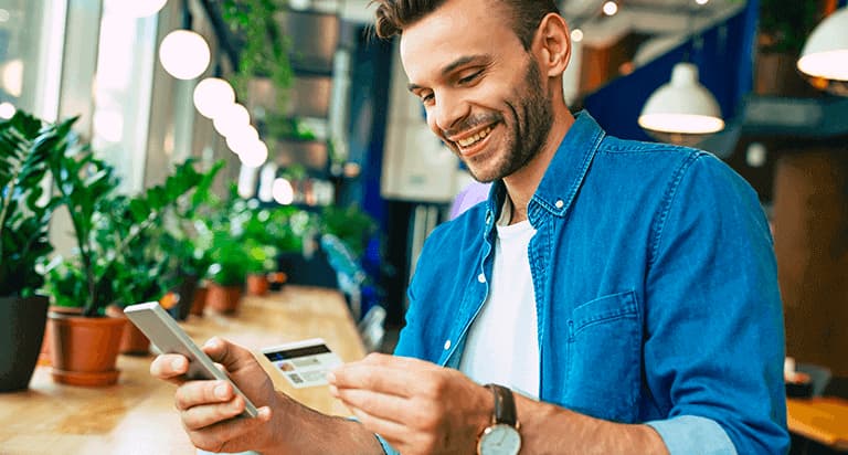 A man smiling down at the credit card in his left hand and the cellphone in his right hand