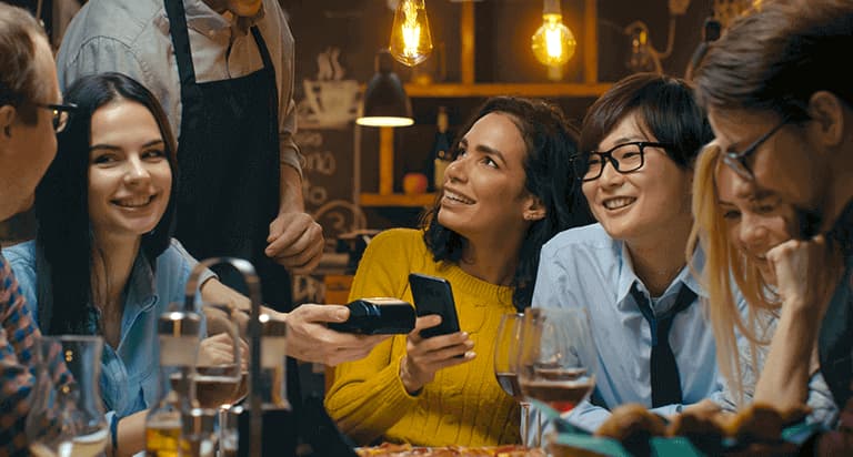 A group of six people smiling and looking at each other while a waiter hands the woman in the middle the payment terminal