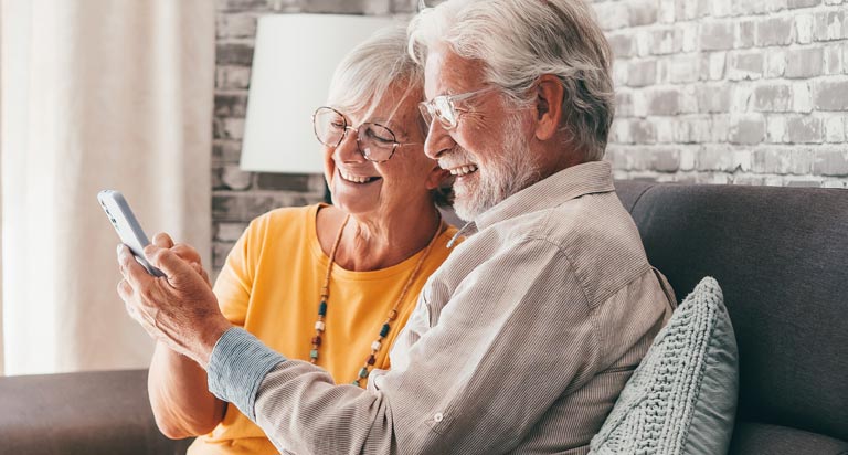 An elderly couple sit on a couch in a brightly lit room with big smiles on their faces while they look at a cell phone.