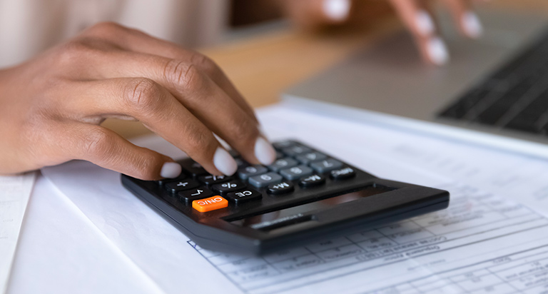 A person sits at a desk while using a calculator and laptop in front of them.