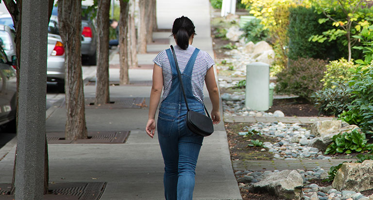 A woman wearing overalls over a striped shirt walks down a sidewalk. She is carrying a black crossover purse. There are parked cards and a row of trees to her left and townhomes to her right.