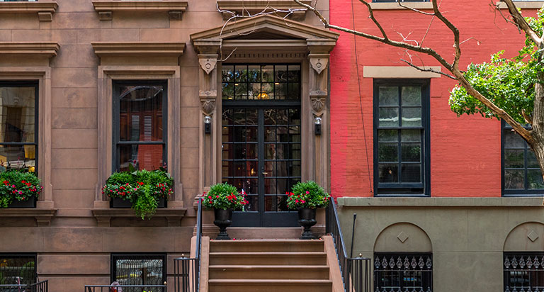 Three rowhouses in a city. The center townhouse has a brown stone exterior and a wrought iron front door with plants on the front steps.