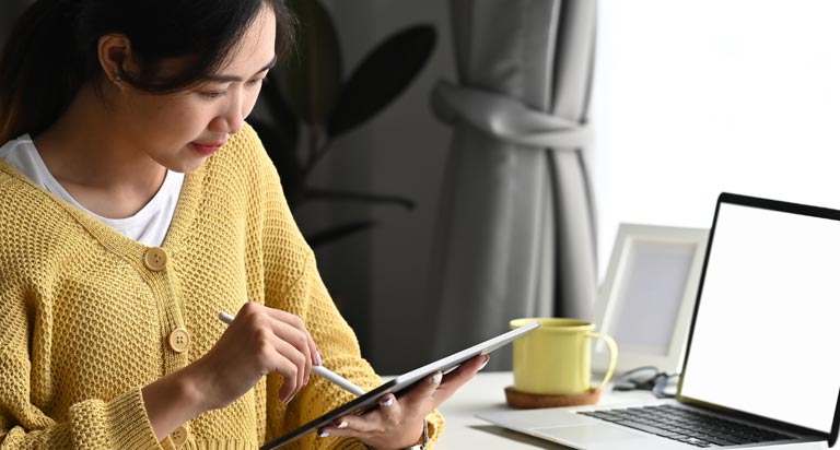 A woman with dark hair and a yellow sweater sits at a desk working on a tablet with a stylus. A laptop rests on the desk.