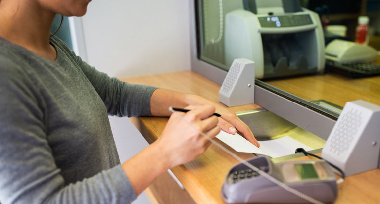 A woman with dark hair and a pen in hand stands at a bank teller window and slides a piece of paper into the transfer slot.