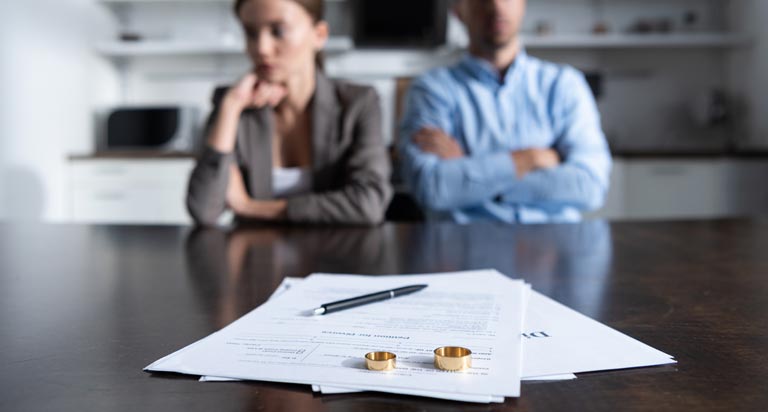 A couple sits at a table in discontent. Divorce documents with wedding rings resting on top sit on the table in front of them.
