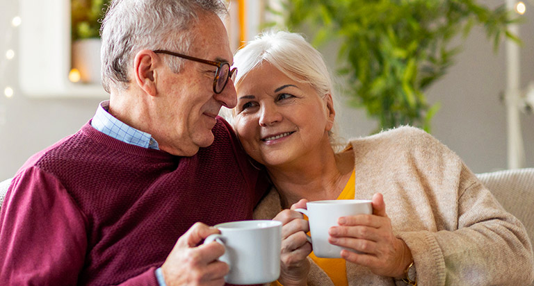 A couple sits together on the couch smiling at each other with white coffee mugs in their hands.