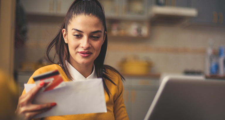 A woman sits at her kitchen table while using her laptop and reading financial papers.