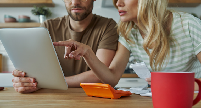 A man and woman sit at a kitchen table. The man is holding a tablet and the woman is pointing at something on the screen.