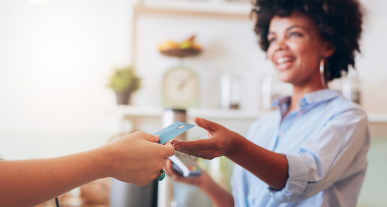 A smiling female cashier reaches over a counter to receive a debit card from a customer who is purchasing a green drink.