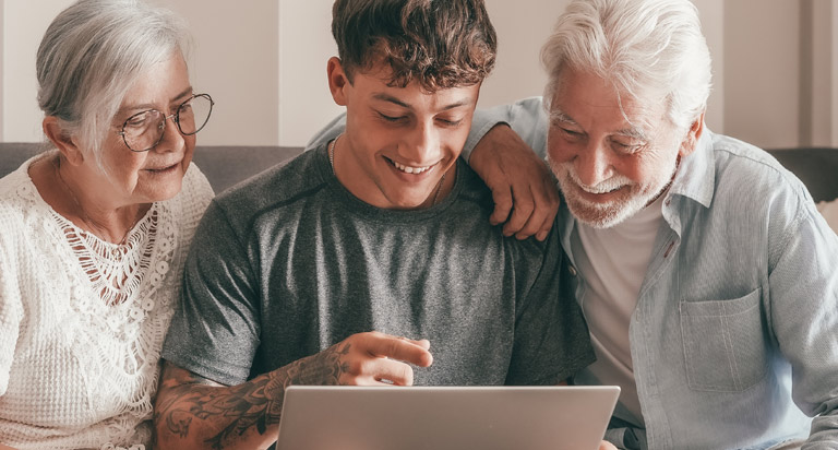 A happy young man and his grandparents sit together on a couch while looking down at a laptop and smiling.