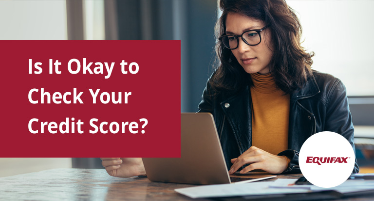 A woman looks down at a laptop on the desk in front of her. The text “Is It Okay to Check Your Credit Score?” is overlaid.