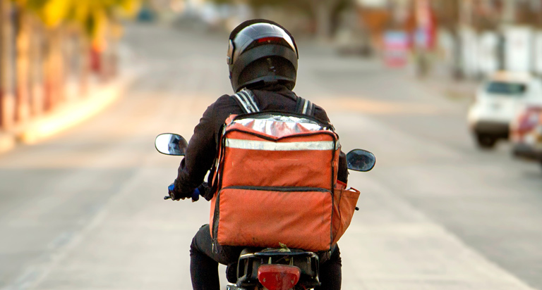 A delivery driver wearing a black helmet on a scooter rides down a street with an insulated bag resting on the back.