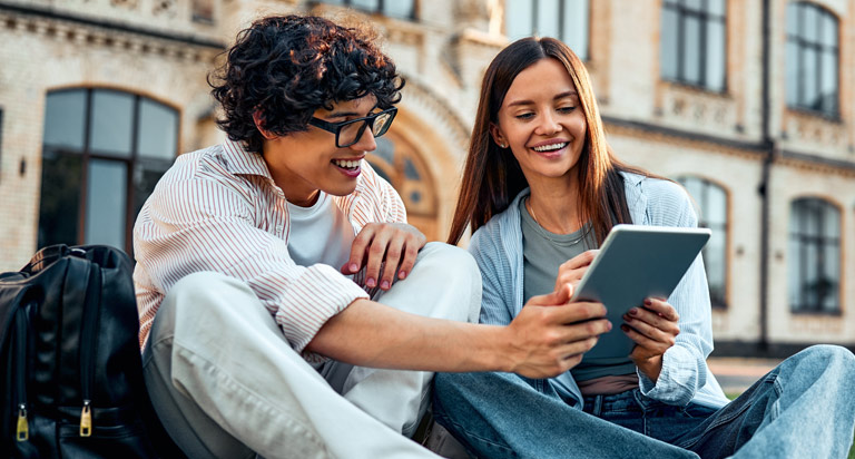 Two college students — one male and one female, sit on a campus lawn. They're smiling, laughing and looking at a tablet.