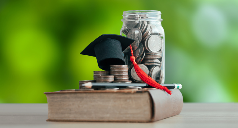 On a wooden surface, a book rests on its side with a jar of change, a pencil, and a small graduation cap sitting on it.