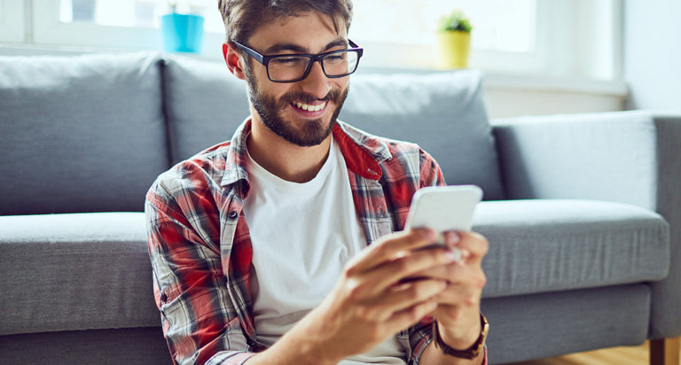 A smiling man with glasses, a beard and short brown hair looks down at his mobile phone as he sits on the floor.