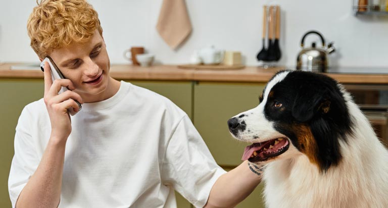 A young man with red hair and a white t-shirt is talking on the phone while petting a dog with black and white fur.