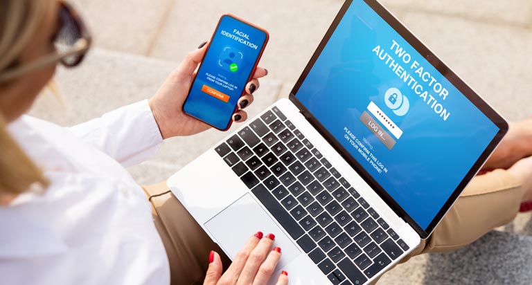 A woman with blonde hair sits with a laptop and mobile phone in hand. Both devices show a multi-factor authentication screen.
