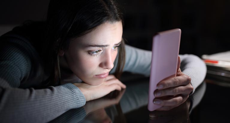 A dark-haired young woman rests her head on a dark surface while looking at a cell phone with a sad look on her face.