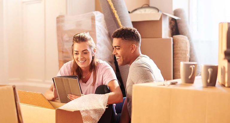 A young man and woman sit on the floor surrounded by moving boxes. They both smile as they look at a tablet together.