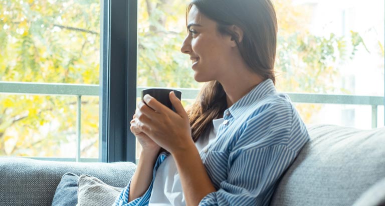 A woman with long brown hair sits on a couch smiling with a cup of coffee in her hands as she looks out the window.