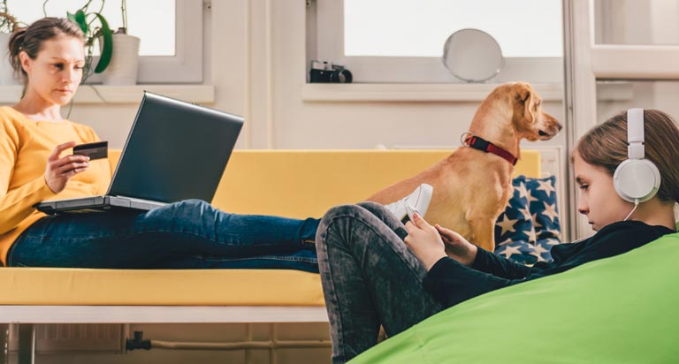 A woman lays on a couch with her laptop and a credit card. A young girl is looking at her phone and a dog sits next to her.