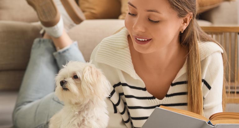 A young woman lays on the floor with her feet up and a book in her hands. She looks over at a small dog next to her.