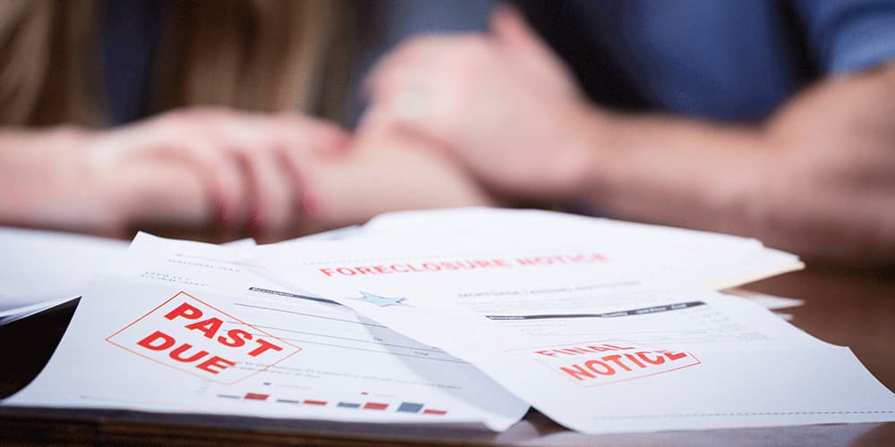 Two people sit in front of a pile of apers scattered on a table with PAST DUE, FINAL NOTICE, and FORECLOSURE stamped on them
